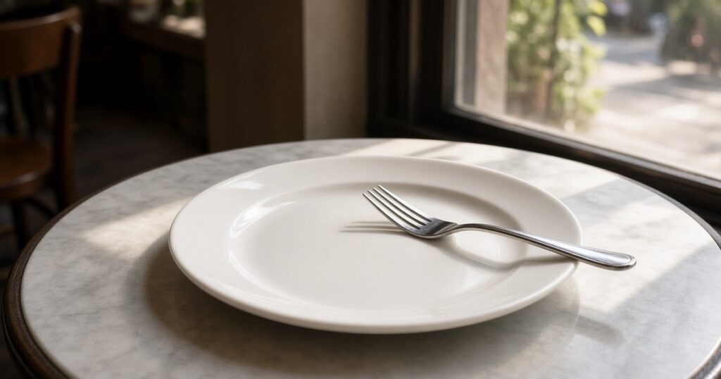 An empty white plate and fork on a cafe table by a window.